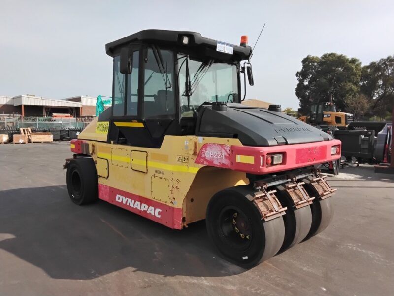 Yellow and pink Dynapac road roller with three metal drums, parked in an industrial lot; used for soil and asphalt compaction.