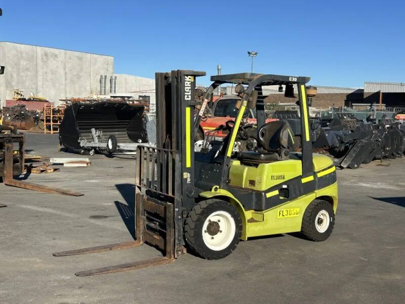Green Clark forklift with extended forks sits in an outdoor industrial yard among metal scraps and equipment, ready to lift loads.
