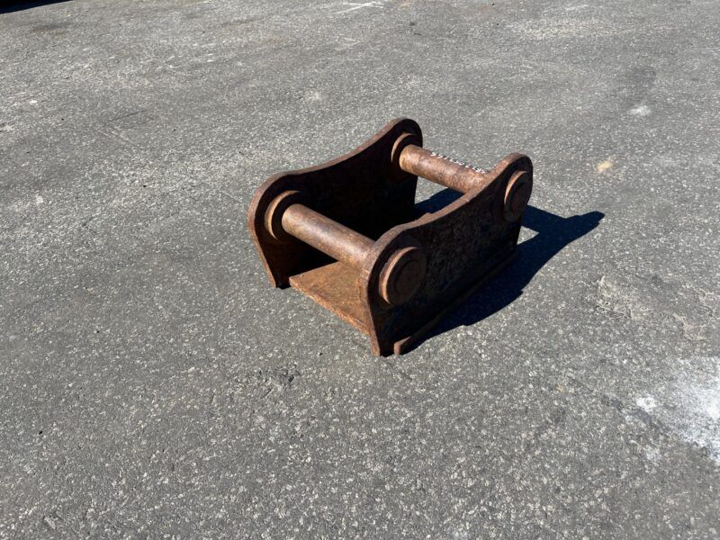 Rusty metal two-pin coupler attachment lying on asphalt, likely for heavy machinery.