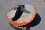 Rusty blue circular industrial cutter head with orange rust on a paved surface, viewed from above.