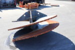 Rusty metal industrial blade or scoop resting on asphalt in an outdoor yard with pallets and cones in the background.