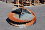 Close-up of a large rusted circular metal cutting wheel on an asphalt surface with orange industrial equipment in the background.