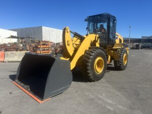 Yellow front-end loader with a large gray bucket parked on an industrial lot, a worker visible in the cab.