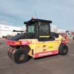 Yellow and red Dynapac road roller parked in an industrial yard with a warehouse in the background.