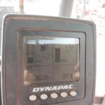 Close-up of a Dynapac digital instrument panel on a dusty surface, showing gauge bars and numeric readouts such as hours and percentages