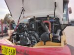 Open-hood view of a heavy-duty truck engine bay with hoses, pumps, and radiator exposed; yellow body with pink rear and license plate H101668 visible