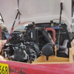 Open-hood view of a heavy-duty truck engine bay with hoses, pumps, and radiator exposed; yellow body with pink rear and license plate H101668 visible