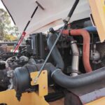Close-up of a diesel engine in a yellow heavy machine, showing hoses, a radiator fan, and hydraulic lines inside the open engine bay.