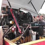 Close-up of an industrial engine with hoses, valves and cables under an open hood; red and yellow frame in the foreground.