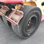 Large road roller with three steel drums and attached sweeping brushes for cleaning a paved surface.
