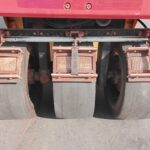 Rear view of a three-drum road roller with large smooth steel drums and rusted metal scrapers.