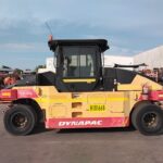 Yellow Dynapac CP224 road roller parked on asphalt in an industrial yard, with stacked equipment in the background.