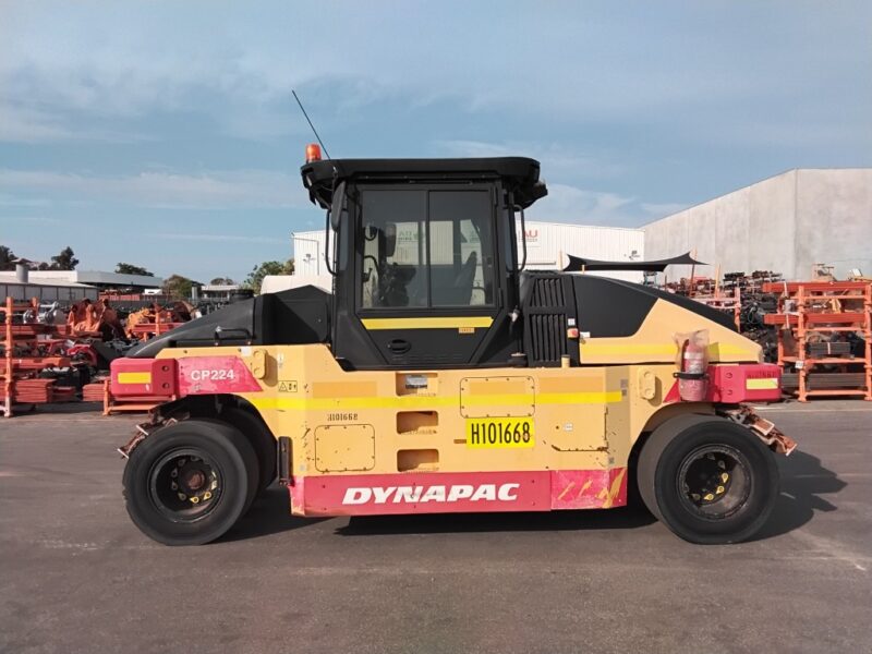 Yellow Dynapac CP224 road roller parked on asphalt in an industrial yard, with stacked equipment in the background.