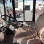 Interior of a dusty tractor cab with steering wheel and joystick; outside is a yard of orange construction equipment.