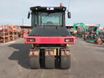 Front view of a red and black industrial road roller in a yard with machinery and stacked materials in the background.