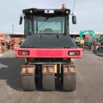 Front view of a red and black industrial road roller in a yard with machinery and stacked materials in the background.