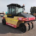 Yellow and pink Dynapac road roller with three metal drums, parked in an industrial lot; used for soil and asphalt compaction.