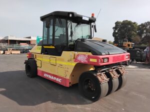 Yellow and pink Dynapac road roller with three metal drums, parked in an industrial lot; used for soil and asphalt compaction.