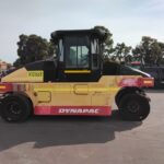 A Dynapac road roller with a yellow body and red base parked on a paved lot, single large front drum and an enclosed cab for the operator.