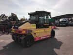 Yellow and pink Dynapac road roller in an industrial yard with trucks in the background.