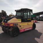Yellow and pink Dynapac road roller in an industrial yard with trucks in the background.