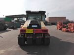 Rear view of a heavy road roller parked in an industrial yard, with orange racks to the right and storage containers in the background.