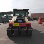 Rear view of a heavy road roller parked in an industrial yard, with orange racks to the right and storage containers in the background.