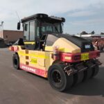 Front-right view of a yellow and red Dynapac road roller with a black enclosed cab parked in an industrial yard.