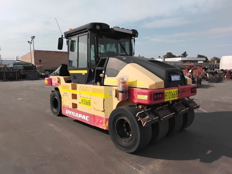 Front-right view of a yellow and red Dynapac road roller with a black enclosed cab parked in an industrial yard.