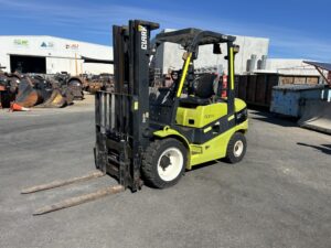 Green CLARK forklift parked in an outdoor industrial yard with rusted attachment pins and a blue sky above the warehouse.