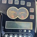 Dashboard of a machine with two circular gauges, a digital display, and a row of function icons around the edges, including seatbelt and parking brake indicators.