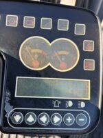 Dashboard of a machine with two circular gauges, a digital display, and a row of function icons around the edges, including seatbelt and parking brake indicators.