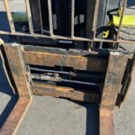 Rusty forklift fork assembly with extended tines resting on asphalt; hydraulic components and frame visible in a rugged, outdoor setting