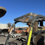 Rusty forklift parked in an outdoor industrial yard with blue sky and other machinery in the background.