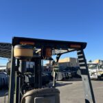 Forklift in an outdoor industrial yard with trucks and equipment under a clear blue sky.