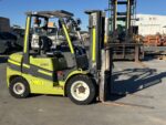 Green and black forklift parked in an industrial yard with forks extended to the right and equipment in the background.