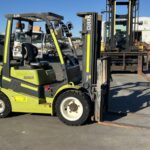 Green and black forklift parked in an industrial yard with forks extended to the right and equipment in the background.