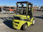 Green forklift in an outdoor industrial yard, with other heavy machinery and trailers in the background.