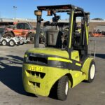Green forklift in an outdoor industrial yard, with other heavy machinery and trailers in the background.