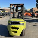 Front view of a green forklift parked in an outdoor industrial yard with orange machinery and metal parts in the background.