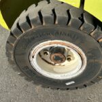 Close-up of a worn forklift tire mounted on a white rim with rust, set against a yellow forklift body on asphalt pavement.