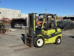 Green Clark forklift with extended forks sits in an outdoor industrial yard among metal scraps and equipment, ready to lift loads.
