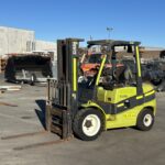 Green Clark forklift with extended forks sits in an outdoor industrial yard among metal scraps and equipment, ready to lift loads.