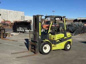 Green Clark forklift with extended forks sits in an outdoor industrial yard among metal scraps and equipment, ready to lift loads.