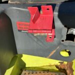 Red battery isolator switch mounted on a dark panel of machinery; label reads 'BATTERY ISOLATOR' with a hinge and mounting holes nearby.