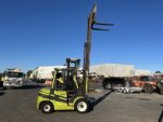 Green and black forklift with extended forks lifting high in an outdoor industrial yard under a clear blue sky.