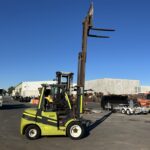 Green and black forklift with extended forks lifting high in an outdoor industrial yard under a clear blue sky.