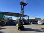 Green forklift in a sunny outdoor industrial yard with metal scrap, containers, and a white truck in the background.