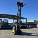 Green forklift in a sunny outdoor industrial yard with metal scrap, containers, and a white truck in the background.