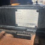 Close-up of a metal identification plate on a black engine, showing model, output, displacement, and engine number, with a weathered label and a rusty surface beneath.